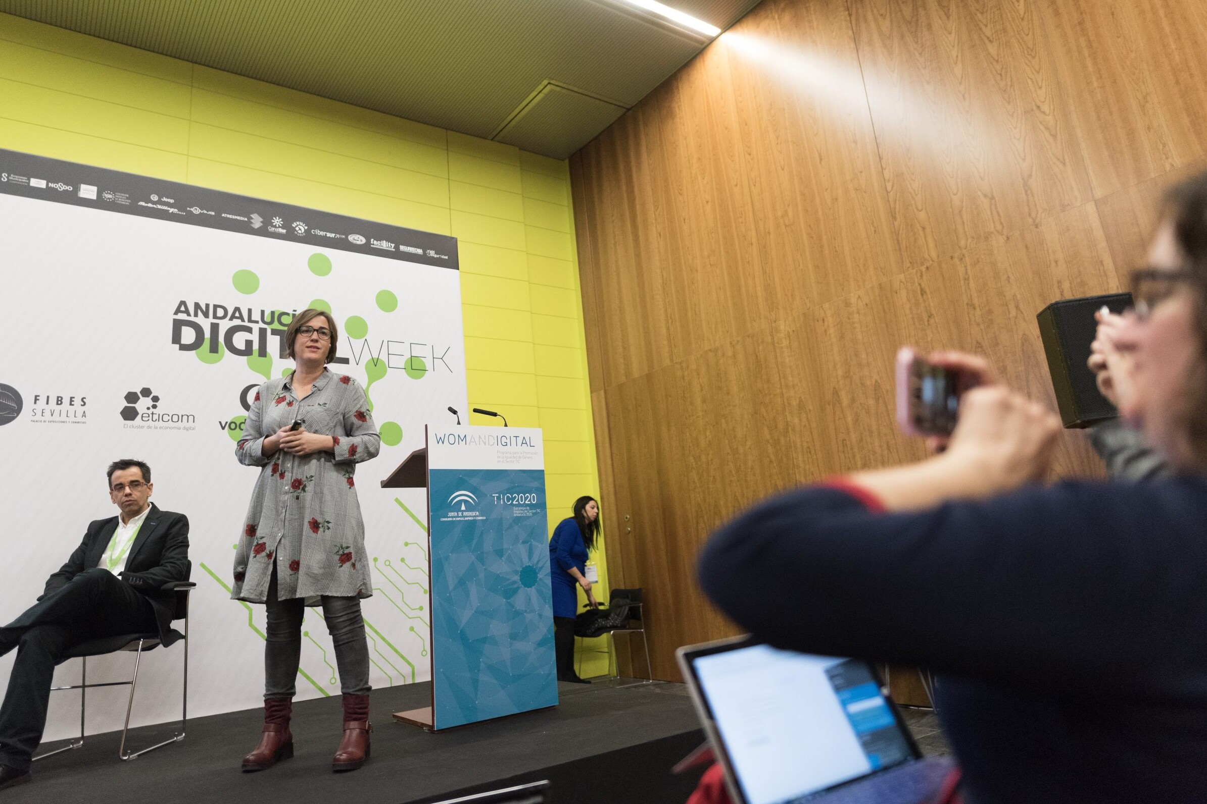 En un escenario, una mujer está hablando mientras varias personas le hacen fotos con desde dispositivos móviles