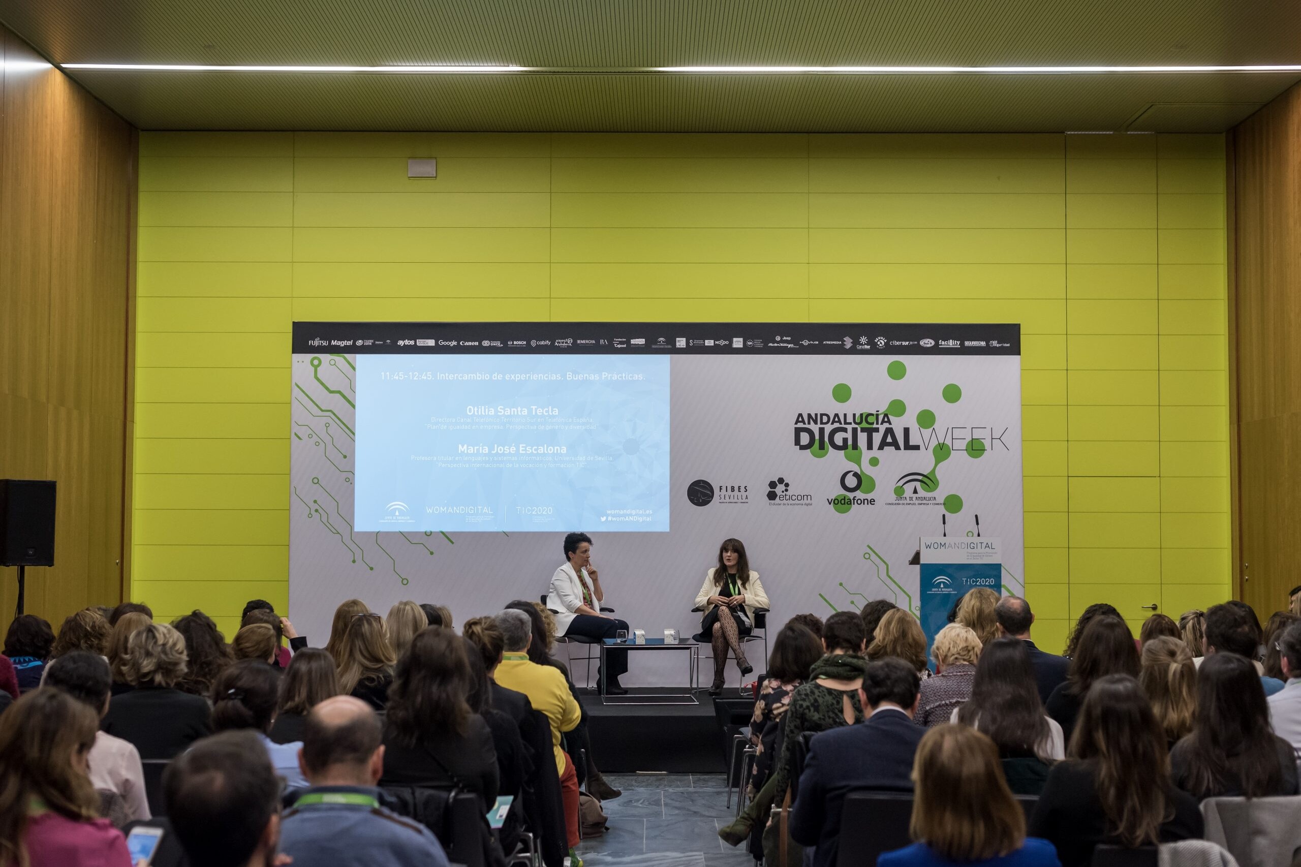 En una sala de conferencias, varias personas sentadas escuchan a dos mujeres dando una conferencia