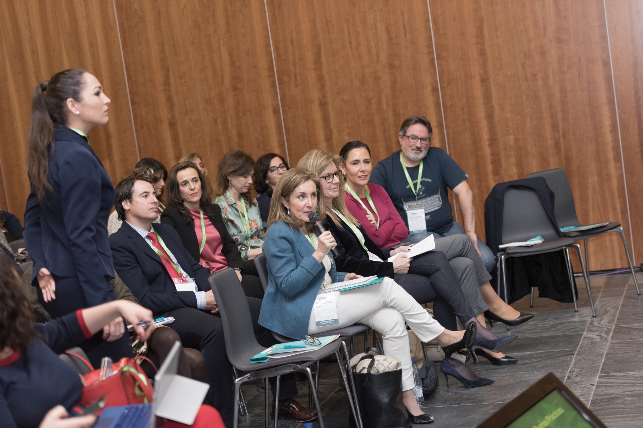 De entre varias personas sentadas en una conferencia, una mujer está de pie hablando