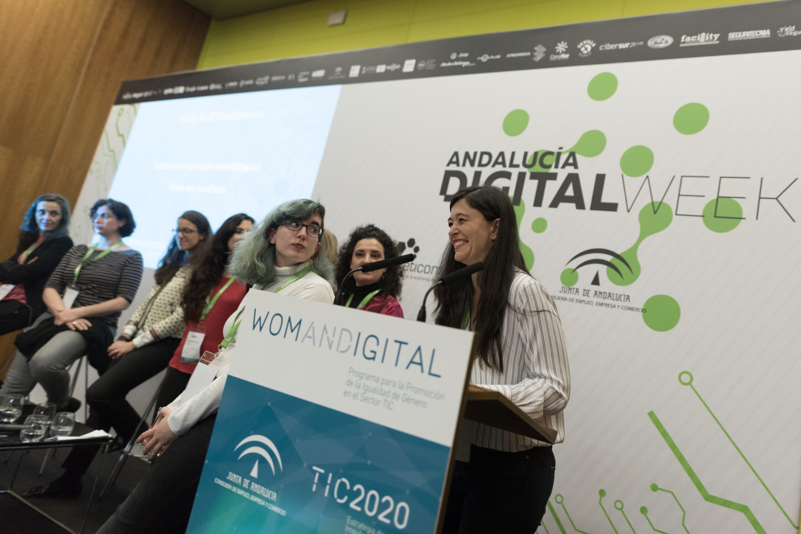 Mujer habla desde el atril del evento junto con otras mujeres en el escenario