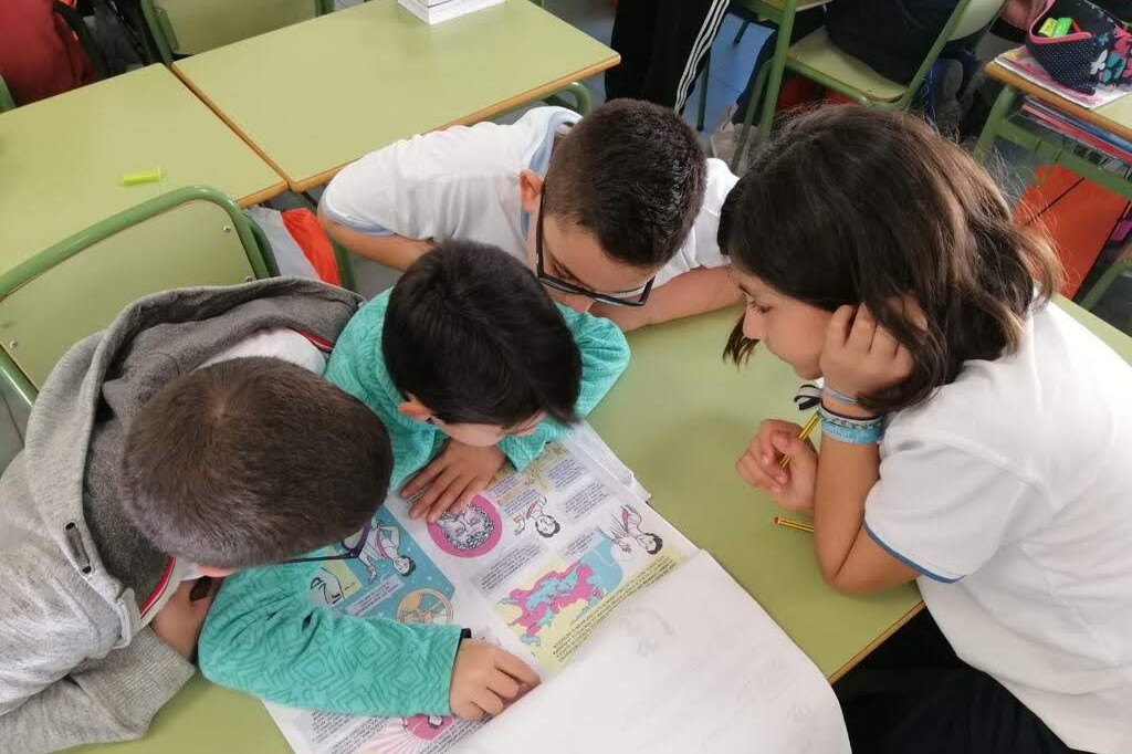 En el pupitre de un aula, tres niños y una niña observan un libro sobre mujeres relevantes en la historia de la ciencia y tecnología