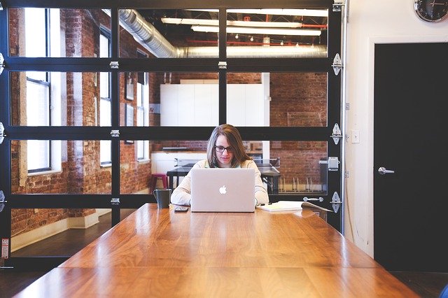 Una mujer está sentada de frente en una mesa de una oficina, trabajando con un portátil